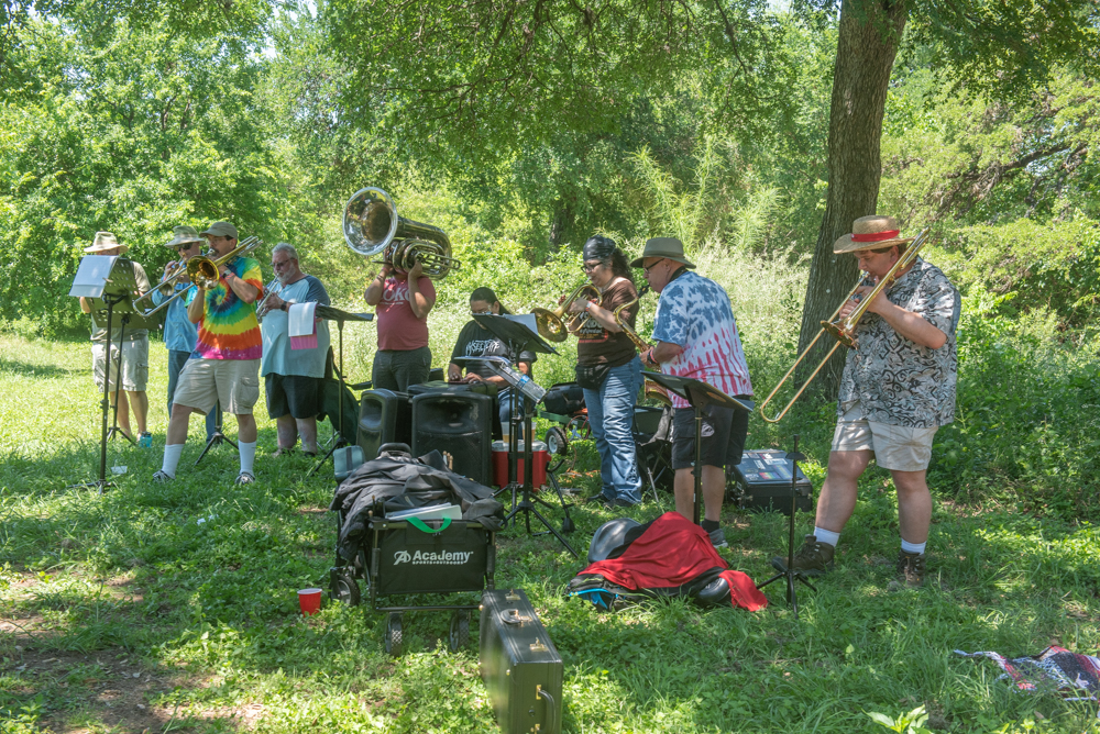 A brass band performs outdoors under the shade of trees, with musicians playing trombones, tubas, and trumpets on a grassy lawn.