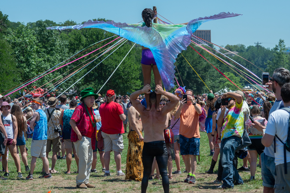 A performer stands on another's shoulders wearing shimmering winged fabric, surrounded by a festive crowd near a maypole with colorful ribbons.