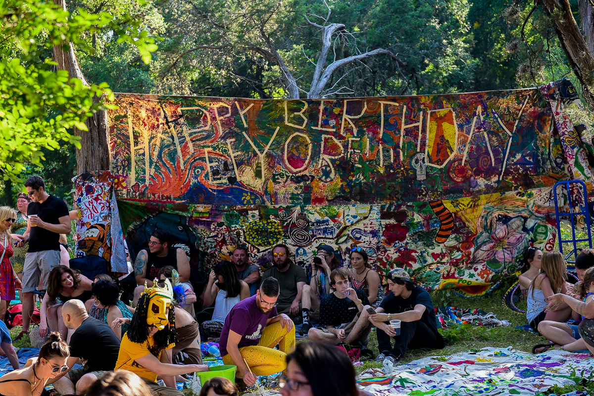 A vibrant "Happy Birthday Eeyore" banner hangs in a wooded area, surrounded by attendees relaxing and painting.