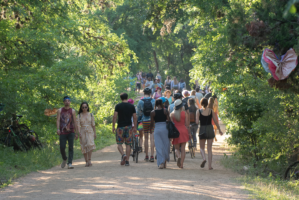 Festival attendees walk along a tree-lined dirt path, some carrying items or pushing bicycles, framed by sunlight.