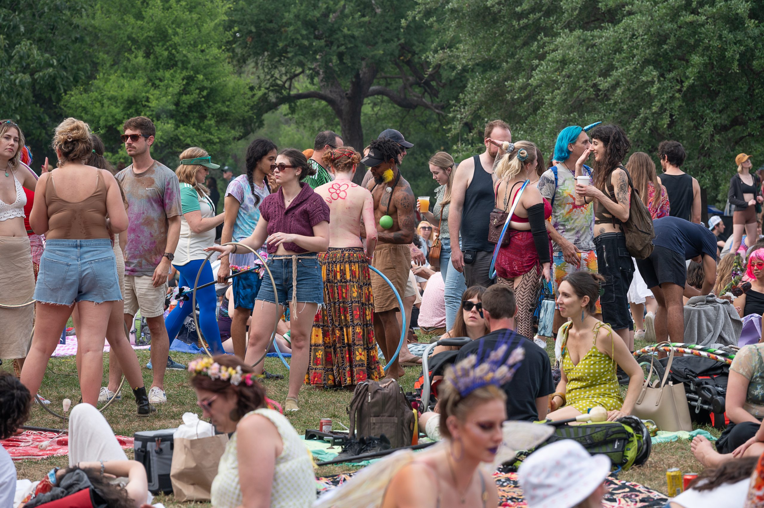 A diverse group of people at a festival relax on blankets, interact, and play with hula hoops on a grassy field surrounded by trees. Some wear colorful and eclectic outfits, including floral crowns and tie-dye clothing, creating a lively and vibrant atmosphere.